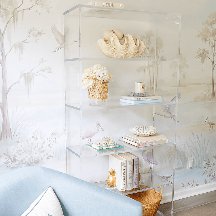 A clear acrylic bookcase with 5 interior shelves decorated with books, candles, decor, and flowers against a floral wallpapered wall.