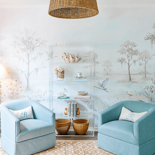 A clear acrylic bookcase with 5 interior shelves decorated with books, candles, decor, and flowers against a floral wallpapered wall between 2 blue lounge chairs.