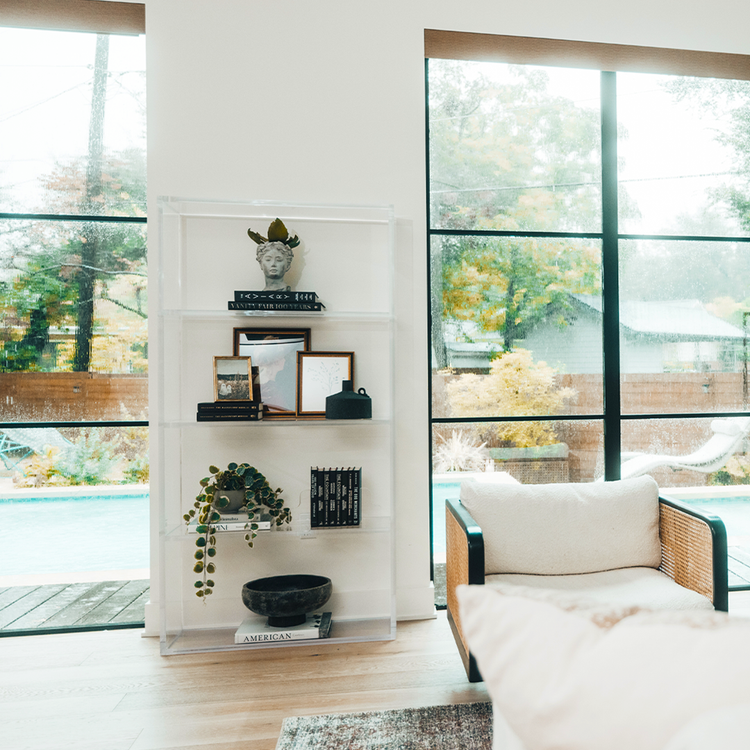 Vertical clear acrylic 4 interior shelf bookcase decorated with plants, vases, books, and decor placed between 2 large picture windows in a living room.