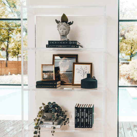 Closeup view of a vertical clear acrylic 4 interior shelf bookcase decorated with plants, vases, books, and decor placed between 2 large picture windows in a living room.