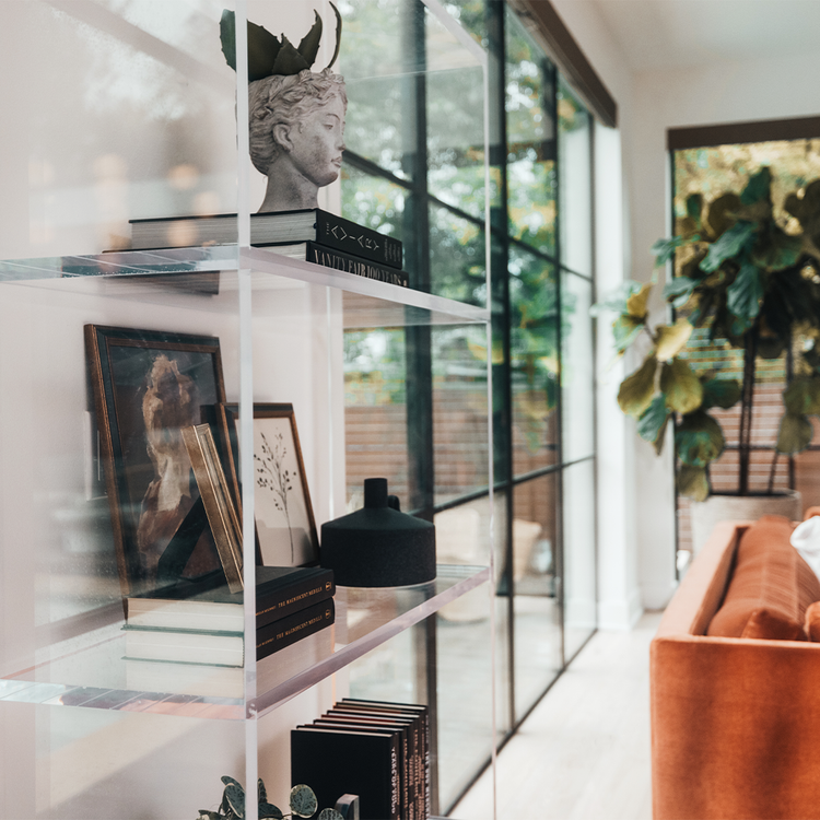 Sideview of a vertical clear acrylic 4 interior shelf bookcase decorated with plants, vases, books, and decor placed between 2 large picture windows in a living room.