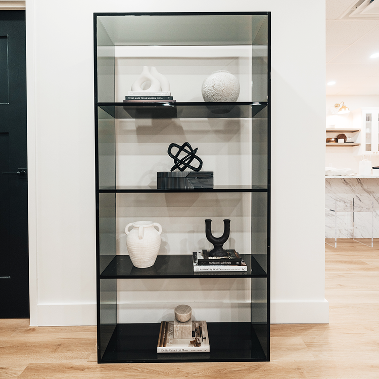 Vertical smoke gray acrylic 4 interior shelf bookcase decorated with vases, books, and decor placed against a white wall in a living room.
