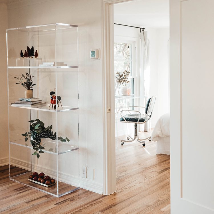 Vertical clear acrylic 5 interior shelf bookcase decorated with plants, vases, books, and decor placed near the entrance of a bedroom also featuring a clear acrylic desk.