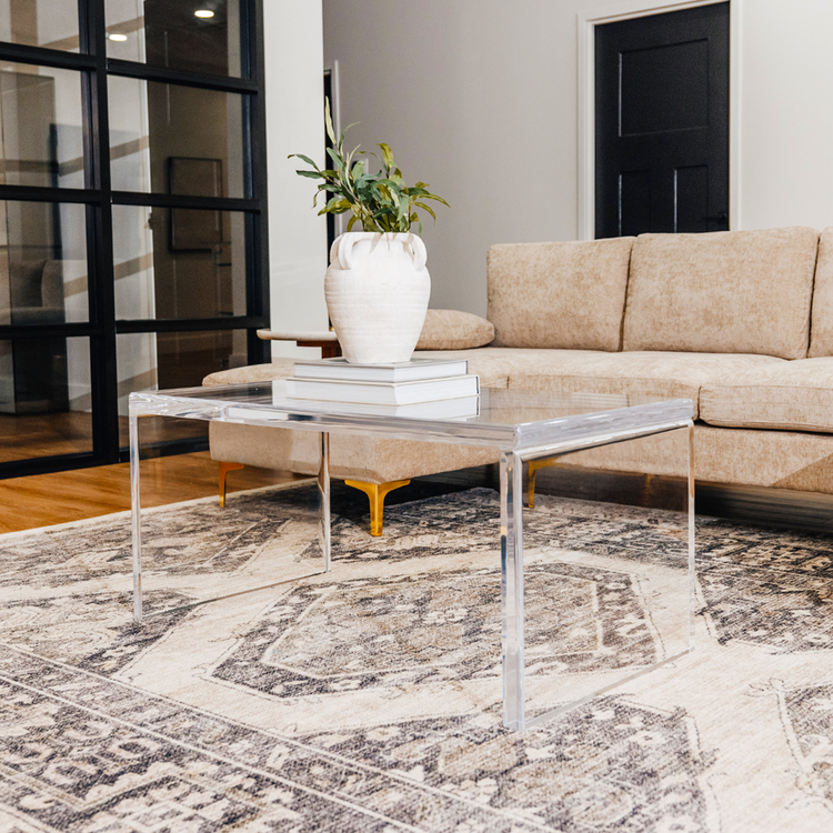 A modern acrylic lucite coffee table placed on a patterned rug, with a white vase on top of it, set against a living room backdrop with a beige couch and black door.