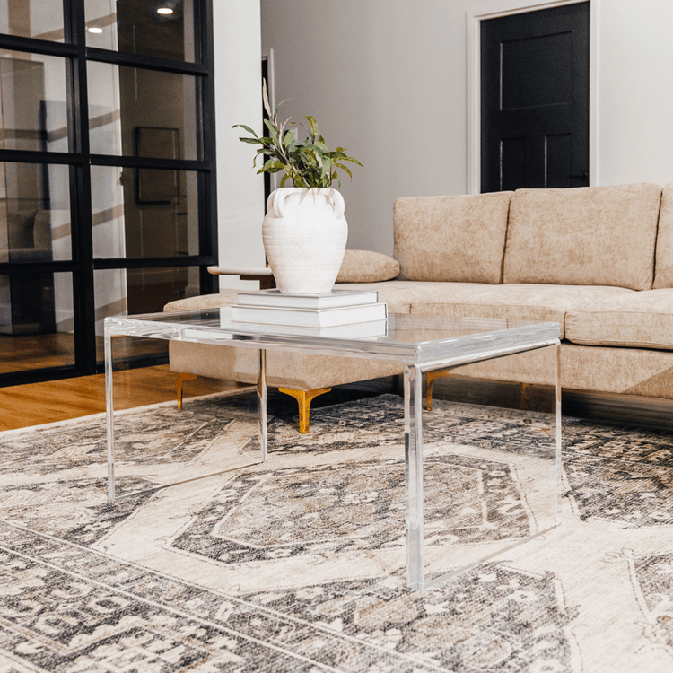 A modern acrylic lucite coffee table placed on a patterned rug, with a white vase on top of it, set against a living room backdrop with a beige couch and black door.