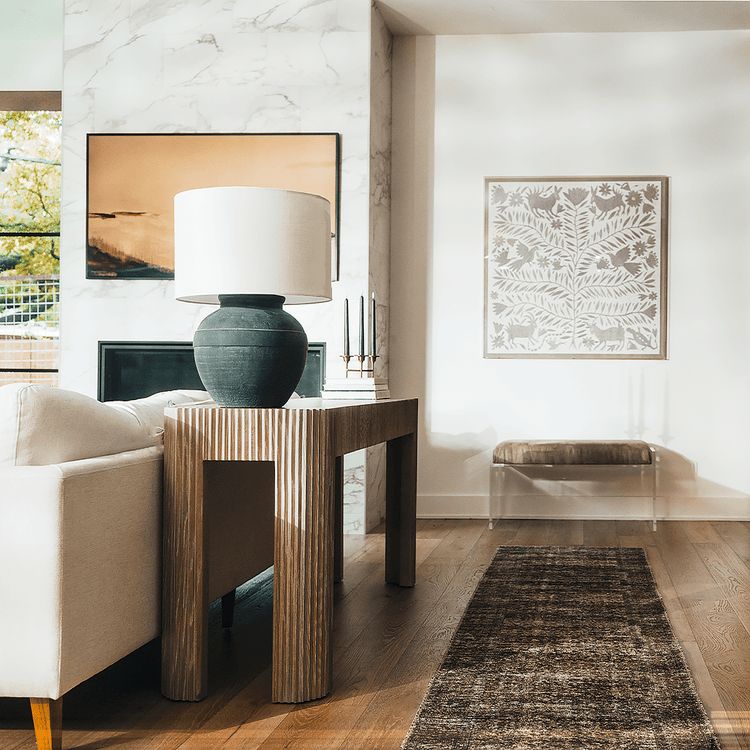 A clear acrylic lucite brown suede upholstered bench placed in a living room as extra seating against a wall with artwork hanging above.