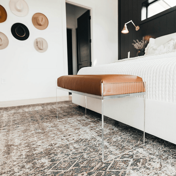 An elegant clear acrylic lucite brown leather upholstered bench at the foot of a bed in a stylishly decorated bedroom setting.