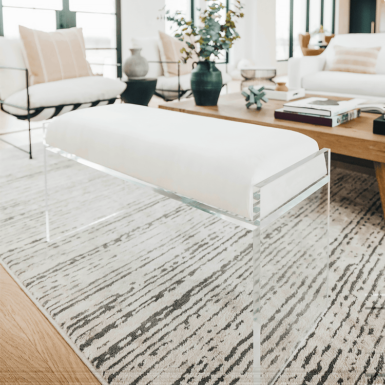 A clear acrylic lucite white velvet upholstered bench placed in a living room as extra seating next to a coffee table, chairs and a couch.