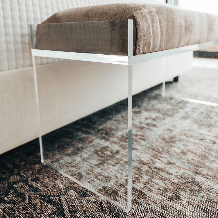 A closeup of a clear acrylic lucite light brown velvet upholstered bench at the foot of a bed in a stylishly decorated bedroom setting.