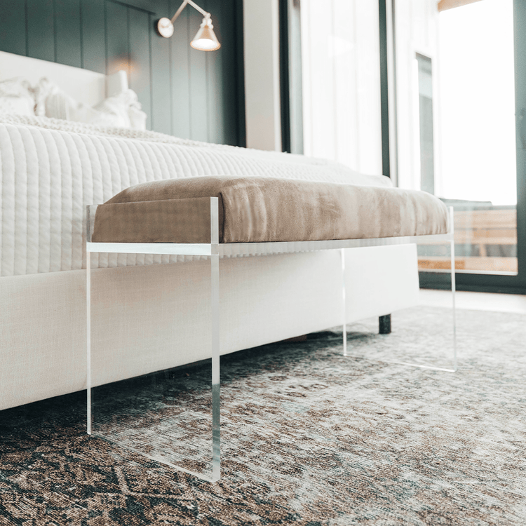 A clear acrylic lucite light brown velvet upholstered bench at the foot of a bed in a stylishly decorated bedroom setting.
