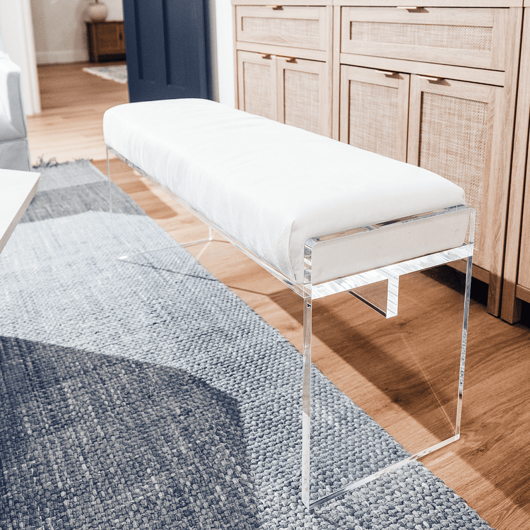 A clear acrylic lucite white velvet upholstered bench placed in a living room as extra seating next to a coffee table and a buffet table.
