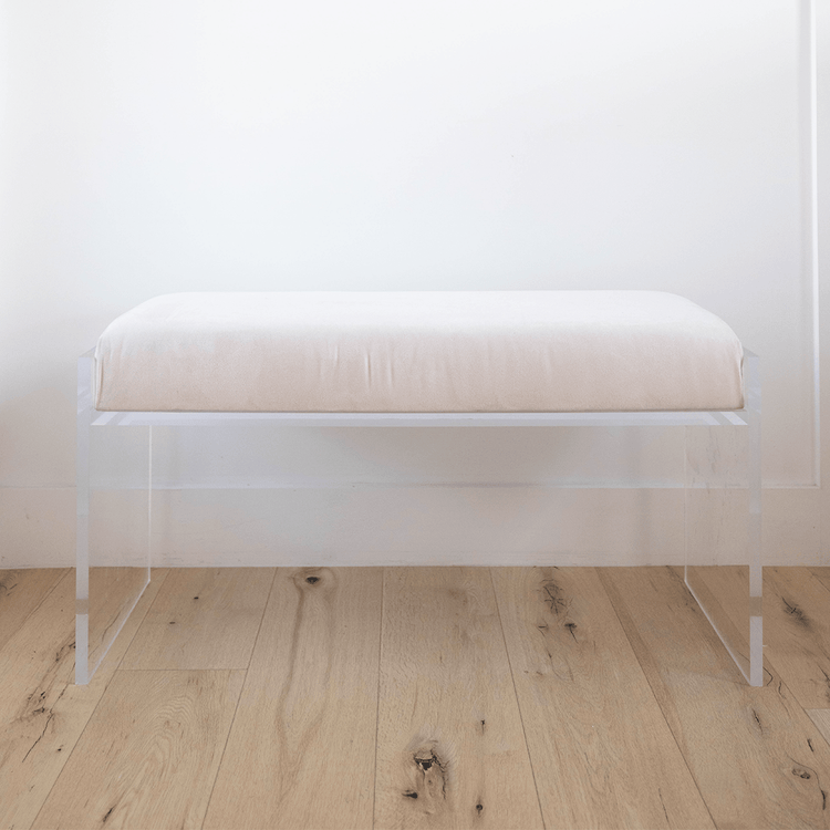 A catalog shot of a clear acrylic white upholstered bench on a wooden floor against a white wall.