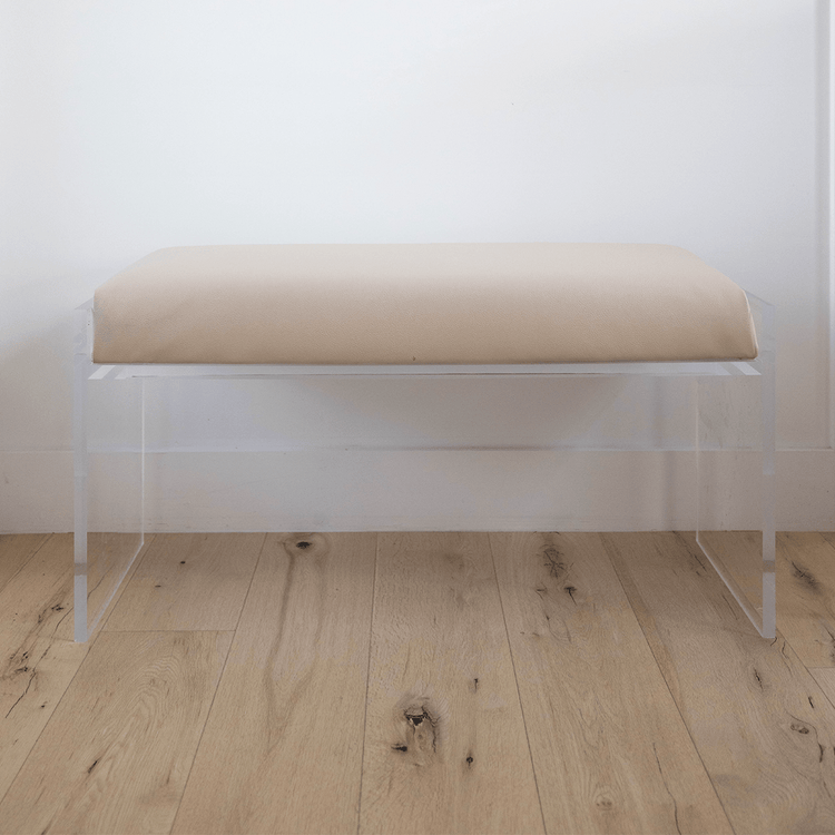 A catalog shot of a clear acrylic beige upholstered bench on a wooden floor against a white wall.