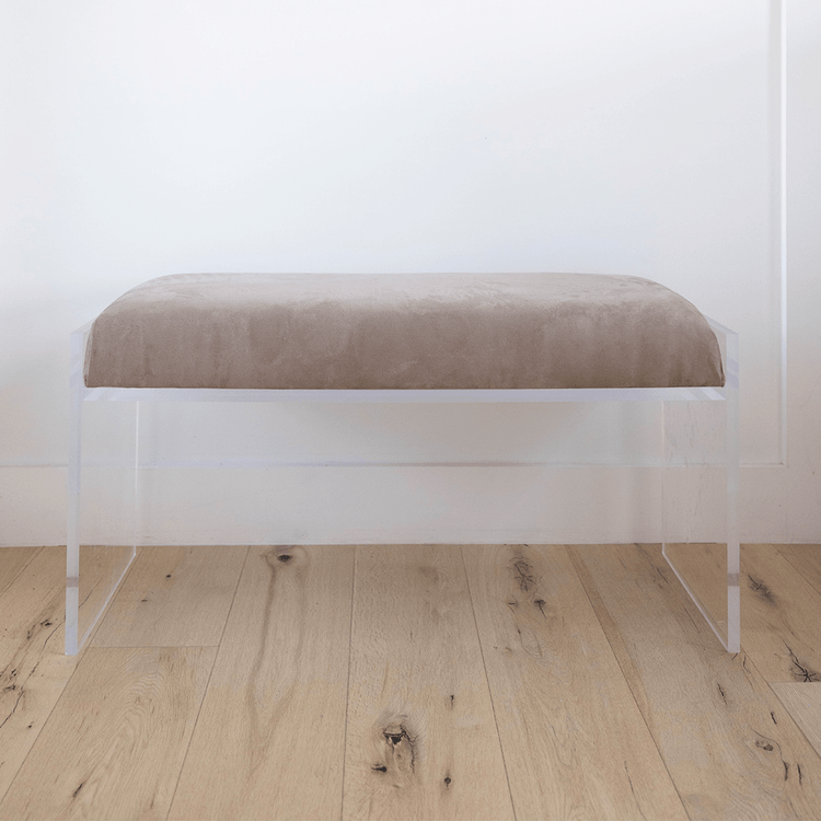 A catalog shot of a clear acrylic brown suede upholstered bench on a wooden floor against a white wall.