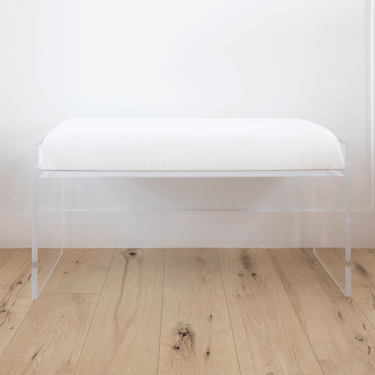 A catalog shot of a clear acrylic white upholstered bench on a wooden floor against a white wall.