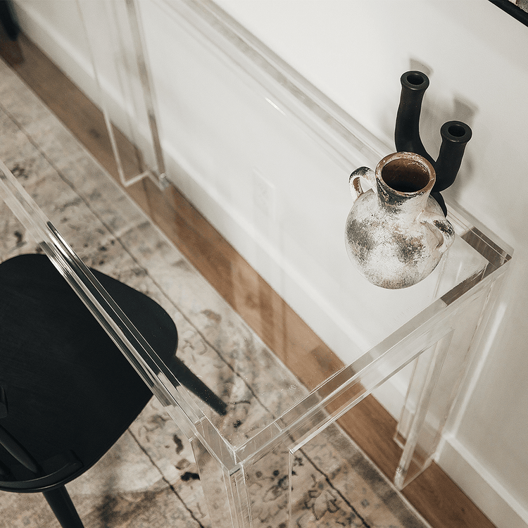 A closeup of the edges of a clear acrylic lucite square leg desk placed against a wall decorated with a vase and candle holder next to a chair.