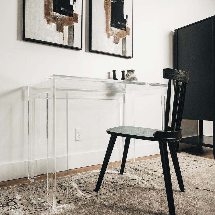 A clear acrylic lucite square leg desk placed against a wall decorated with a vase and candle holder next to a chair.
