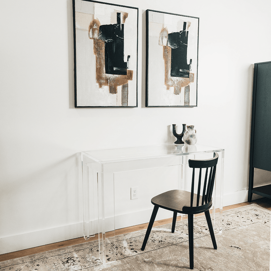 A clear acrylic lucite square leg desk placed against a wall decorated with a vase and candle holder next to a chair.