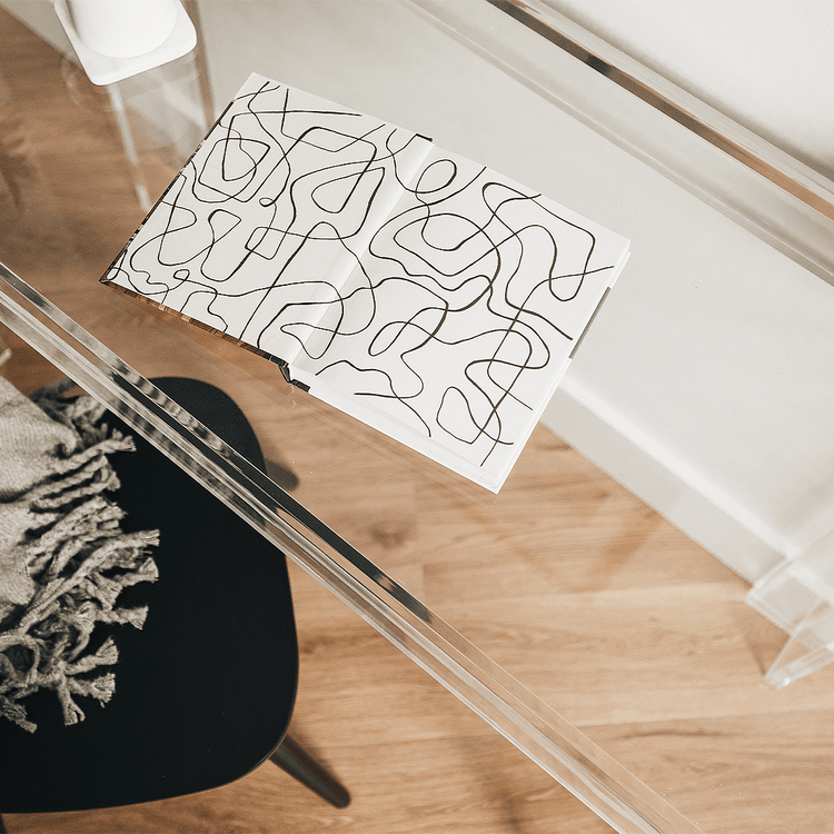 Clear acrylic lucite square leg desk placed against a wall decorated with a coffee cup and a book next to a chair layered with a blanket on the back.