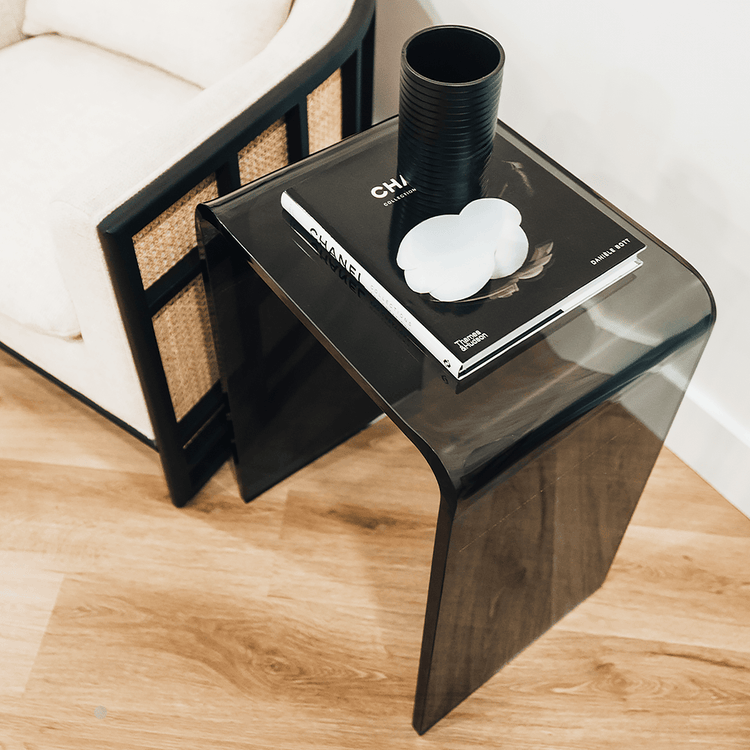 A grey tinted acrylic lucite waterfall edge side table with a book and coffee cup placed next to an easy chair.