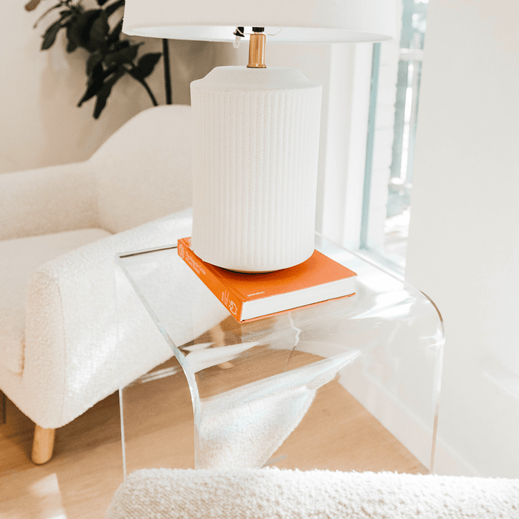 A clear acrylic lucite waterfall edge side table with a lamp and book on top illuminated by sunlight placed against a wall between 2 easy chairs.
