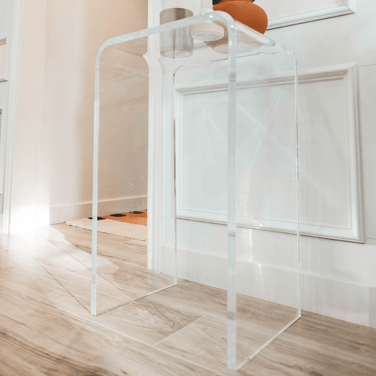 A clear acrylic lucite waterfall edge side table with a candle and decor on top illuminated by sunlight placed against a wall.