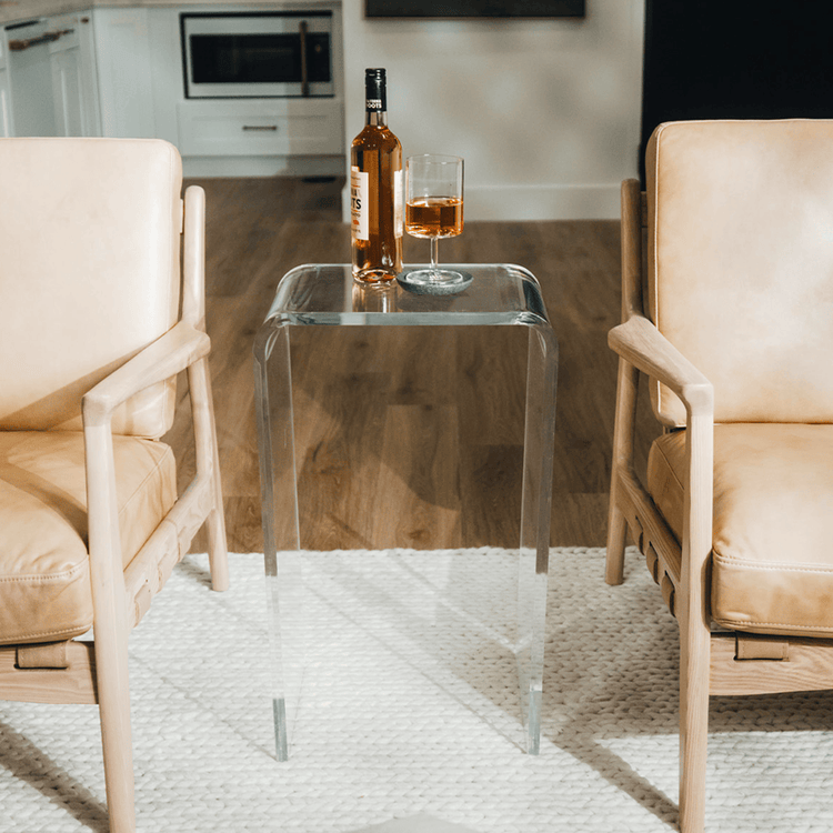 A clear acrylic lucite waterfall edge side table with a bottle of wine and wine glass on top placed between two easy chairs.