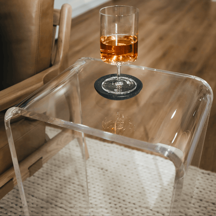A closeup of the edges of a clear acrylic lucite waterfall end table holding a wine glass next to an easy chair.