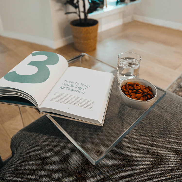 A C-Shaped acrylic lucite side table with a beverage glass, snacks, and a book on top partially slid underneath a sofa in a living room.