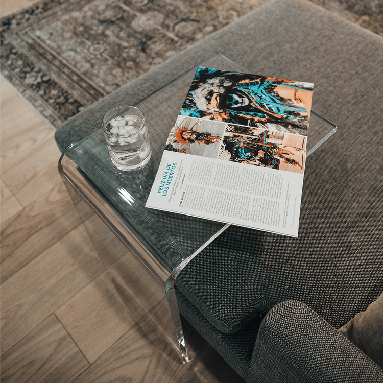 A C-Shaped acrylic lucite side table with a beverage glass and a book on top partially slid underneath a sofa in a living room.