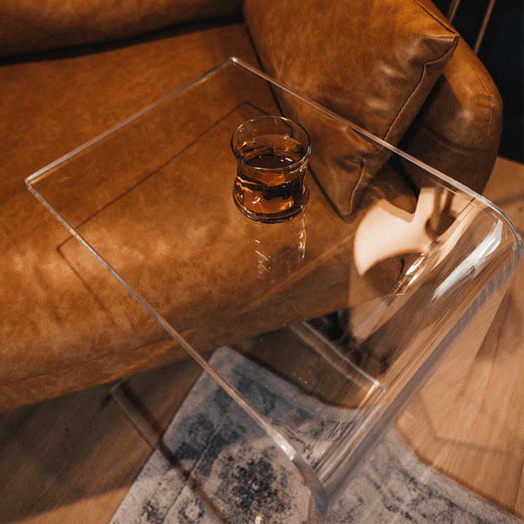 A C-Shaped acrylic lucite side table with a beverage glass on top partially slid underneath a brown leather sofa.