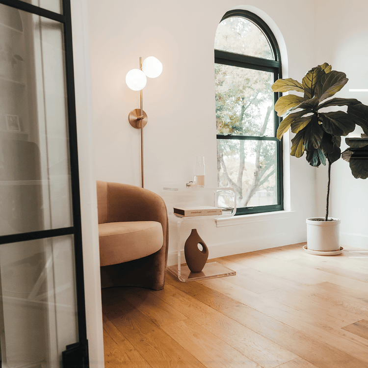 A clear acrylic lucite side table in a modern s-shape with drinks on top and a book on the interior shelf placed beside an easy chair in a living room next to a window and a lamp.