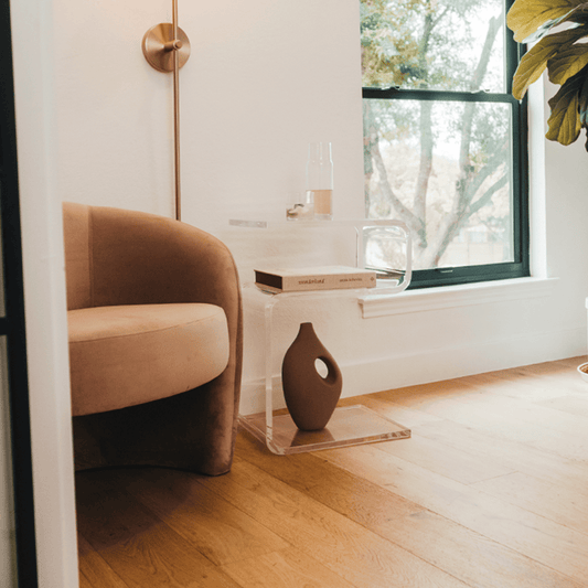 A clear acrylic lucite side table in a modern s-shape with drinks on top and a book on the interior shelf placed beside an easy chair in a living room next to a window and a lamp.