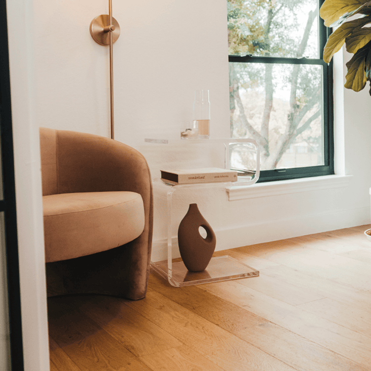 A clear acrylic lucite side table in a modern s-shape with drinks on top and a book on the interior shelf placed beside an easy chair in a living room next to a window and a lamp.