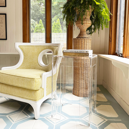 A clear acrylic side table with a square leg style base placed next to a yellow upholstered chair in the corner of a well-lit living room setting.