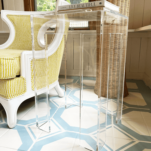 A clear acrylic side table with a square leg style base placed next to a yellow upholstered chair in the corner of a well-lit living room setting.