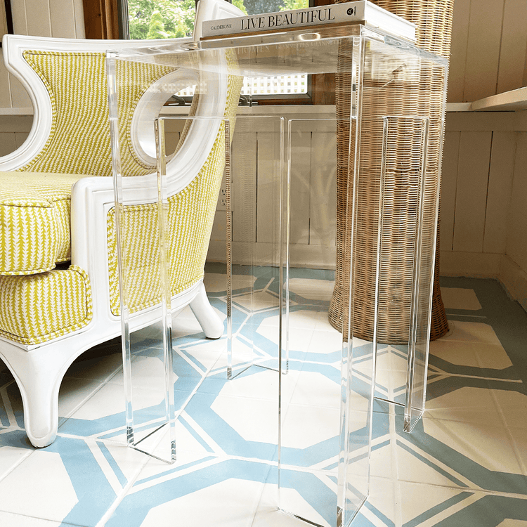 A clear acrylic side table with a square leg style base placed next to a yellow upholstered chair in the corner of a well-lit living room setting.