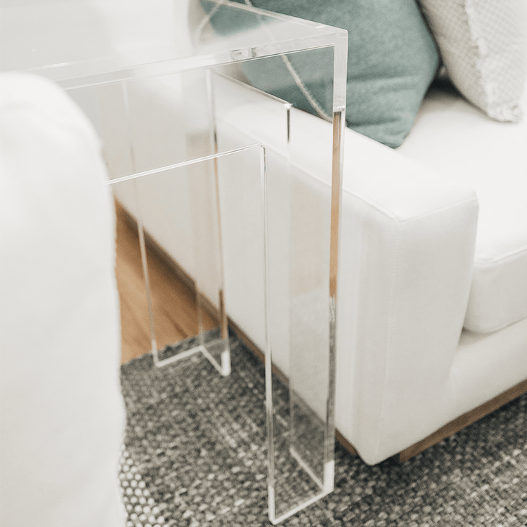 A closeup of a clear acrylic side table with a square leg style base placed between a white couch and a white chair in a modern living room.