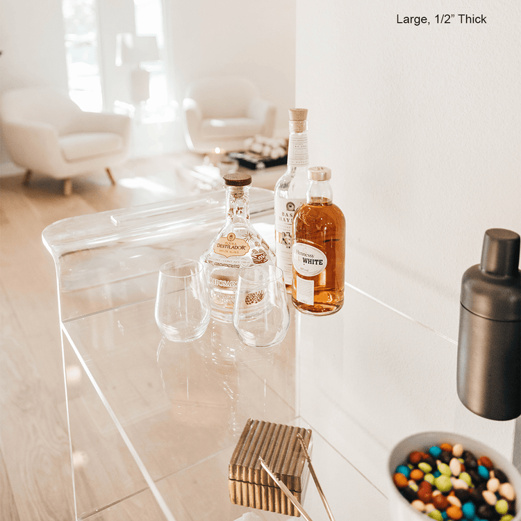 A clear acrylic bar cart featuring 3 shelves, wheels, and clear acrylic handles decorated with bar essentials, candles, and other decor items placed against a living room wall.