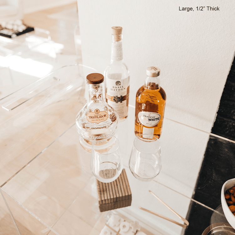 A clear acrylic bar cart featuring 3 shelves, wheels, and clear acrylic handles decorated with bar essentials, candles, and other decor items placed against a living room wall.