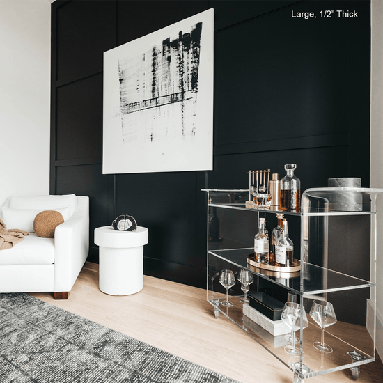 A clear acrylic bar cart featuring 3 shelves, wheels, and clear acrylic handles decorated with bar essentials, candles, and other decor items placed against a living room wall.
