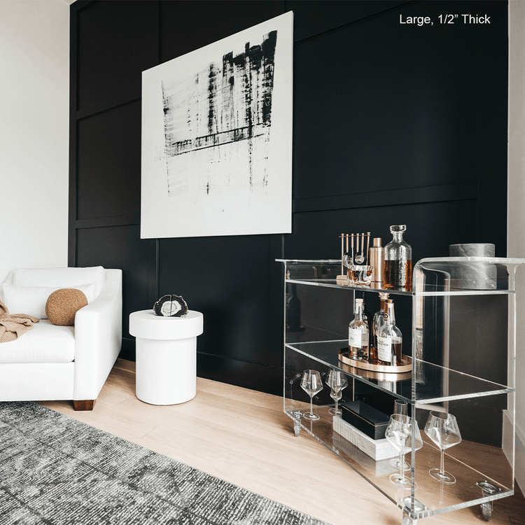 A clear acrylic bar cart featuring 3 shelves, wheels, and clear acrylic handles decorated with bar essentials, candles, and other decor items placed against a living room wall.