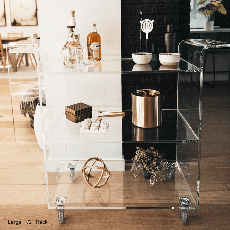 A clear acrylic bar cart featuring 3 shelves, wheels, and clear acrylic handles decorated with bar essentials, candles, and other decor items placed against a living room wall.