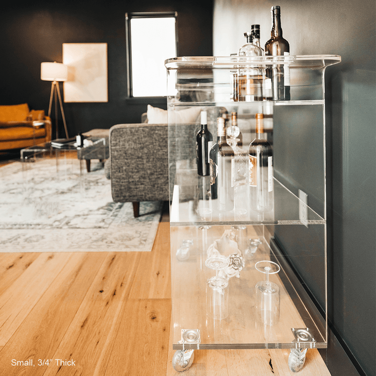 A clear acrylic bar cart featuring 3 shelves, wheels, and clear acrylic handles decorated with bar essentials, candles, and other decor items placed against a living room wall.