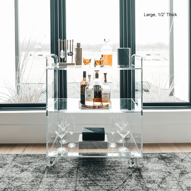 A clear acrylic bar cart featuring 3 shelves, wheels, and clear acrylic handles decorated with bar essentials and glasses in front of a large picture window.