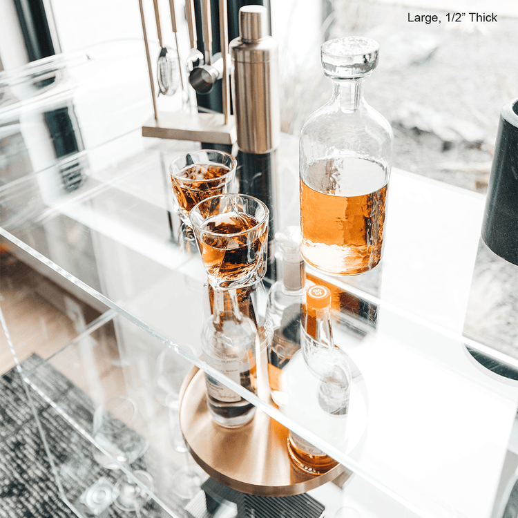 A clear acrylic bar cart featuring 3 shelves, wheels, and clear acrylic handles decorated with bar essentials and glasses in front of a large picture window.
