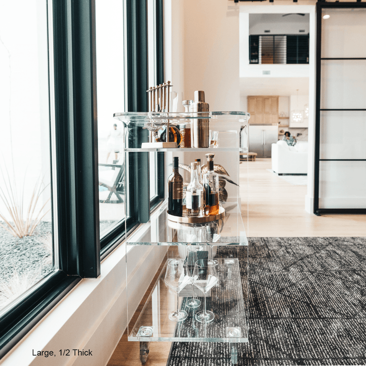 A clear acrylic bar cart featuring 3 shelves, wheels, and clear acrylic handles decorated with bar essentials and glasses in front of a large picture window.