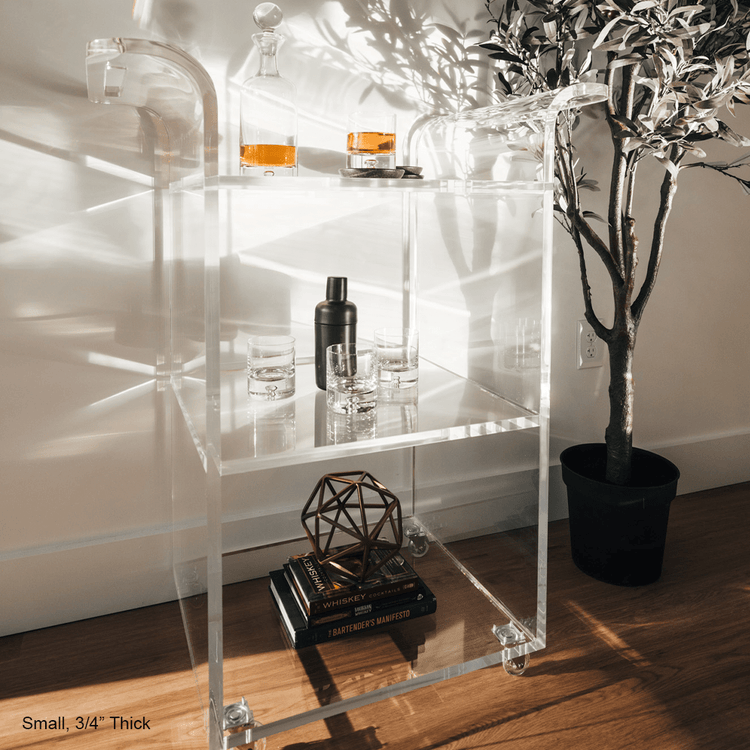 A clear acrylic bar cart featuring 3 shelves, wheels, and clear acrylic handles decorated with bar essentials, candles, and other decor items placed against a living room wall.