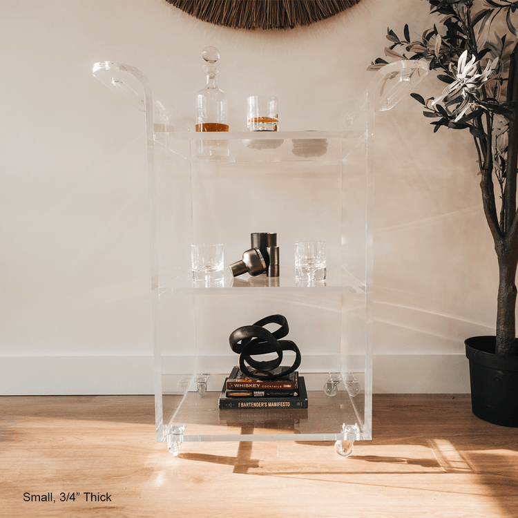 A clear acrylic bar cart featuring 3 shelves, wheels, and clear acrylic handles decorated with bar essentials, candles, and other decor items placed against a living room wall.