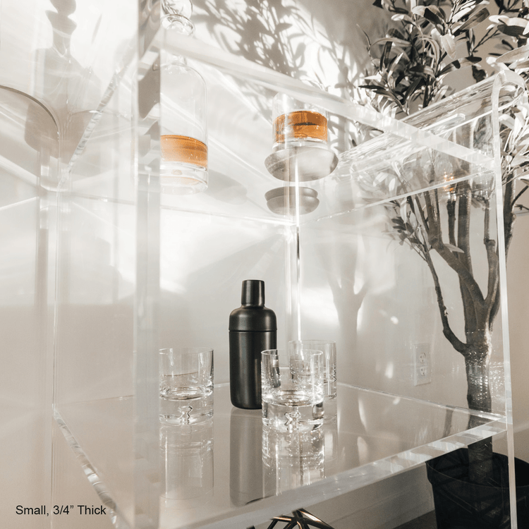 A clear acrylic bar cart featuring 3 shelves, wheels, and clear acrylic handles decorated with bar essentials, candles, and other decor items placed against a living room wall.